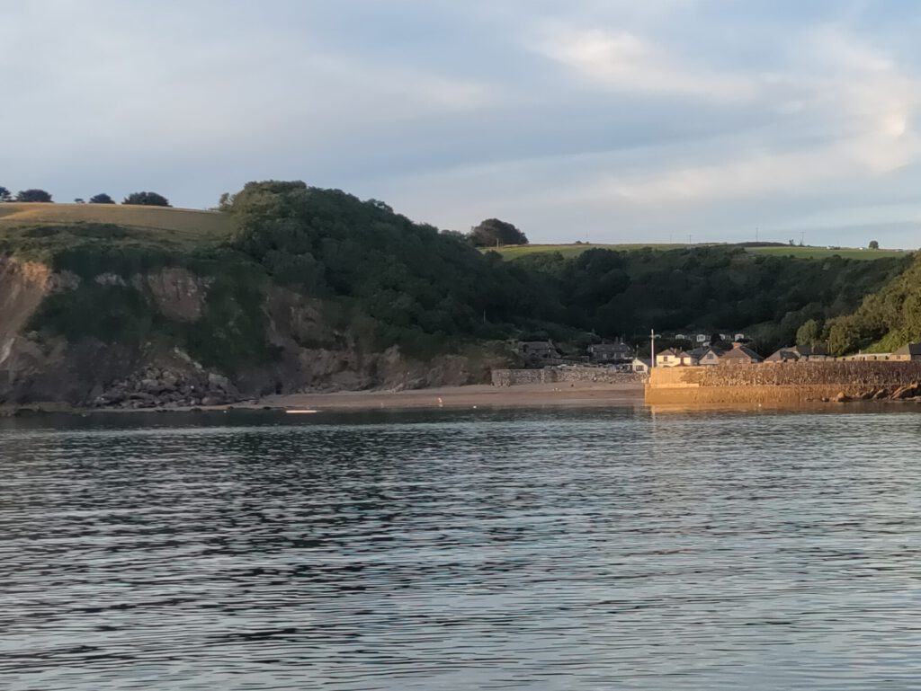 Man blickt vom Meer aus auf  eine Hafenmauer und ein Stück Strand. Dahinter liegen ein paar Häuser in einem Taleinschnitt